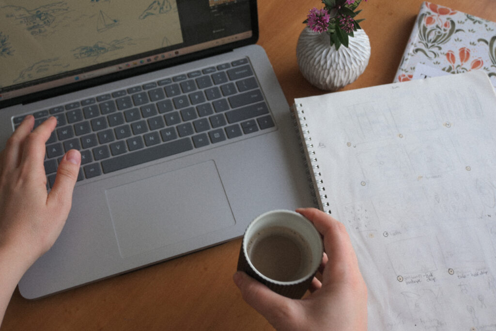 Jenn Kane working at a laptop with sketchbook and coffee, developing repeat pattern concepts in a creative studio workspace