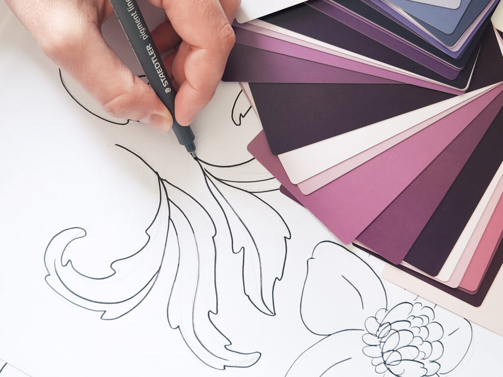Jenn Kane sketching floral surface pattern artwork with a fineliner pen beside a mauve and purple color palette swatch fan