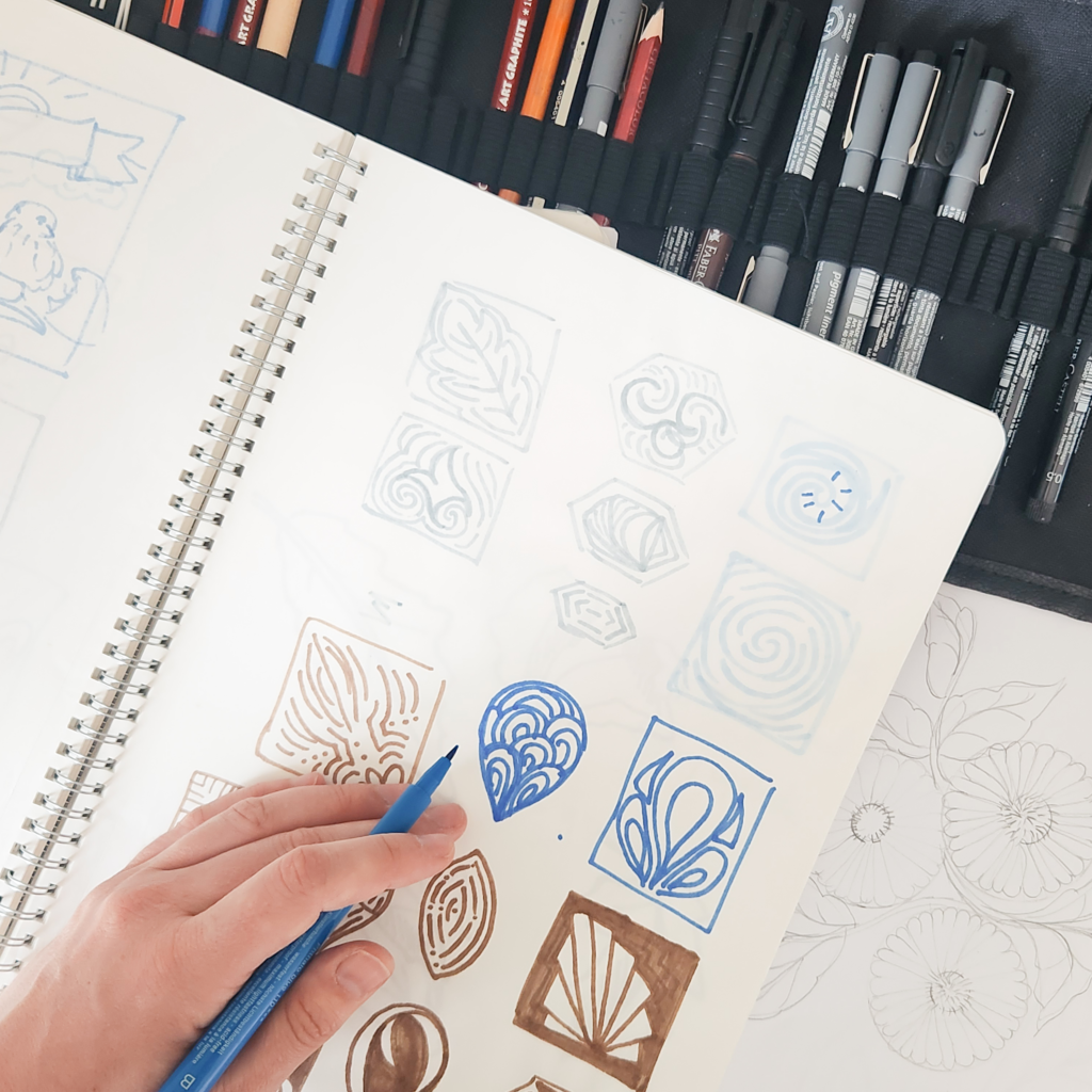 Jenn Kane's hand with marker drawing in a sketch book full of motifs.