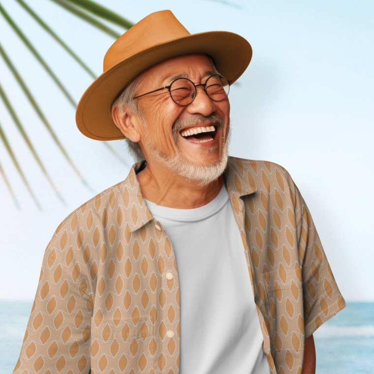 Smiling man in a straw hat wearing a tan short-sleeve shirt with a repeating geometric leaf pattern, standing by the ocean with a palm frond overhead.